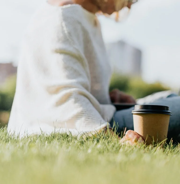 Cup of coffee in woman hands sitting on the grass.